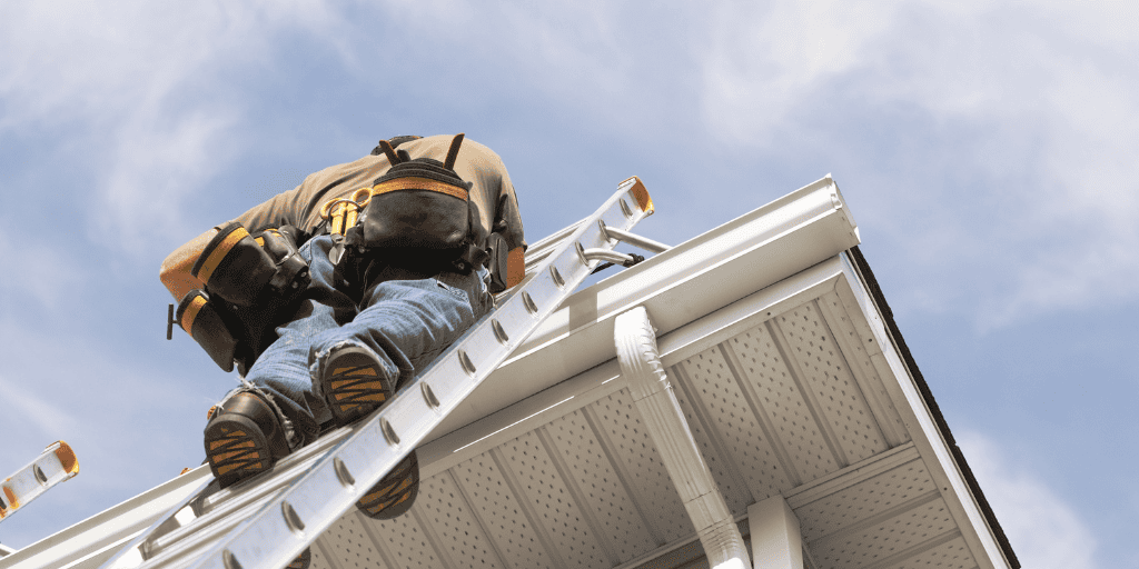 Ladder worker upgrading roof gutter system on house. Alt text: Worker installing or repairing roof gutter on house using ladder, outdoor home maintenance.